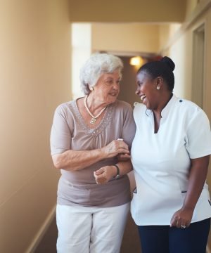 Portrait of happy female caregiver and senior woman walking together at home. Professional caregiver taking care of elderly woman.